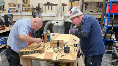 Two men stand either side of a wooden work bench which is covered with various tools. In the background, more tools and materials are seen on the floor and hanging up on the wall