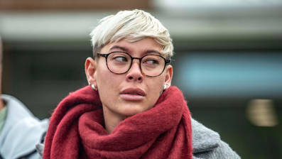 A woman with short blonde hair and glasses, wearing a grey court and red scarf