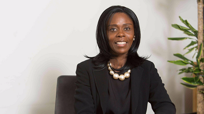 Barbara Mills: A woman sitting in a grey chair. She has black hair and is wearing a black jacket and top, and a white string of pearls.