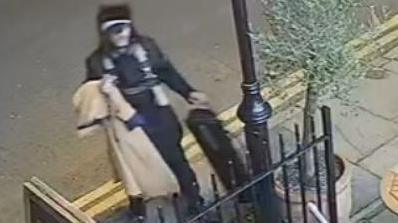 A blurred CCTV image of a man holding a beige trench coat, wearing a black outfit.