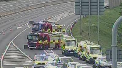 A grainy CC screenshot showing two fire engines and a several ambulances and a police car lined up on a motorway slip road.