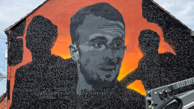 A man painting a black and orange mural. The mural shows a painting of a man looking to the right in black and white on an orange background. T