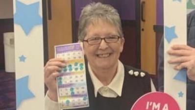 Wendy is looking at the camera and smiling while holding her winning bingo ticket. She is stood behind a cardboard frame that someone is holding in front of her while she has her photo taken. 