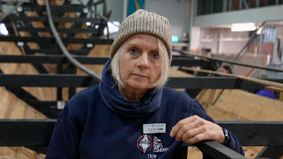 Jacq Barnard stands within the reconstructed ship's hull. One arm rests on black metal framework as she smiles at the camera. She is wearing a grey beanie hat, blue neck snood and a navy jumper with a name badge on her chest.