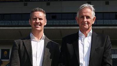 Stephen Vaughan stands next to Worcester Warriors owner Chris Holland in font of the main stand at Sixways. Both are wearing dark suits with white shirts and have their hands crossed in front of their waists. 