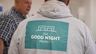 The back of a person who is wearing a grey hoodie with the words The Good Night Project on the back. The individual appears to be talking to a person to their left