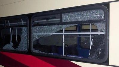 A coach at night in a depot with its windows smashed. There are remnants of shattered glass with large holes in. The coach is cream coloured with red and burgundy stripes on it.