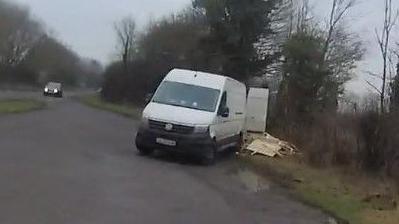 A slightly pixelated image of a white van in a lay-by with a big pile of wood behind it.