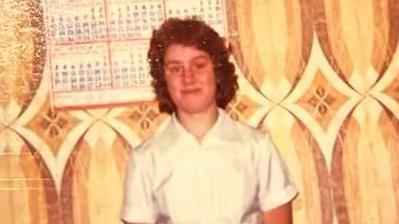 A woman in a white shirt standing in front of wallpaper with a swirling brown 1970s style pattern. The woman has chin-length curly brown hair and is smiling with her mouth closed. A wall calendar is visible behind her.