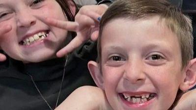 Sienna, nine, who has long brown hair and is wearing a black jumper, smiles at the camera with her fingers in a V-shape, while Brody, seven, smiles while resting his head on his hands.