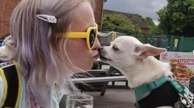 A woman with sunglasses at a pub beer garden table giving a kiss to a chihuahua dog