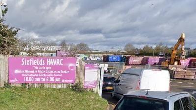 Entrance to Sixfields recycling centre.  There is a purple welcome sign to the right and a cue of traffic through the entrance. A digger is visible to the right and there is a row of shipping containers behind.