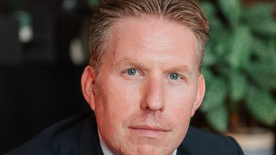 A man looking directly at the camera. He is clean shaven and wearing a dark suit and white shirt.
