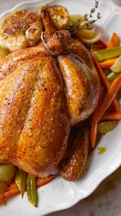 A whole roast chicken sitting on a bed of vegetables on a large white plate. 