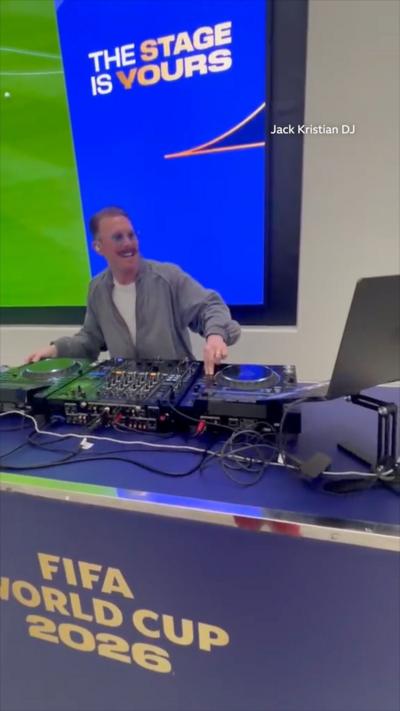 Man wearing a grey jacket, white t-shirt, a moustache and brown hair smiling while Djing. He's standing in front of DJ decks, a big screen behind him with the colour blue dominating. A