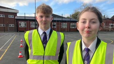 Two children standing in a playground wearing hi-vis jackets over their uniforms with some cones on the ground in the background.