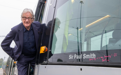 Paul Smith, wearing a dark blue suit, standing in the open door of a tram which has Sir Paul Smith written in the window