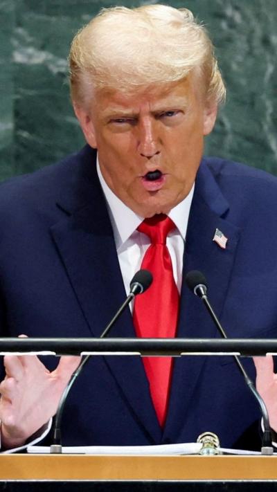 Trump in a dark suit with red tie with two microphones in front of him