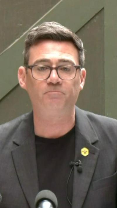 Andy Burnham wearing a black top and suit with a yellow pin and glasses.