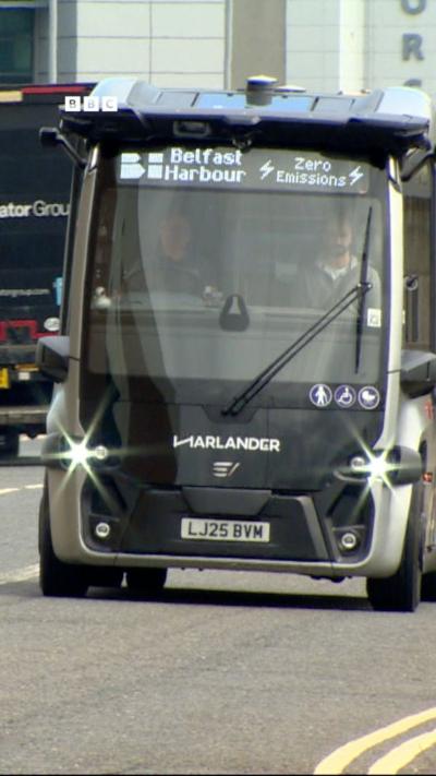 A small silver and black bus with tinted windows on a road. Two people are visible inside. The bus destination says Belfast Harbour. Harlander is along the front of the bus.