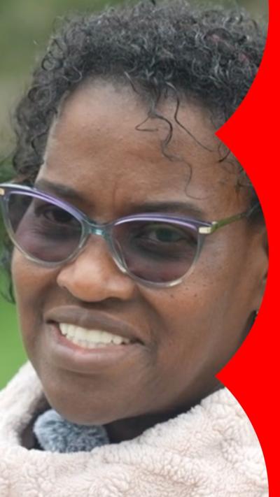 A woman with short, dark curly hair speaks to the camera. She wears sunglasses and a white jumper.