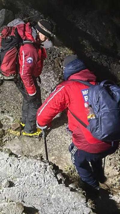Two mountain rescue volunteers walk on the side of a craggy mountain in the dark, wearing headtorches. They are wearing red mountain rescue branded jackets and carrying rucksacks.