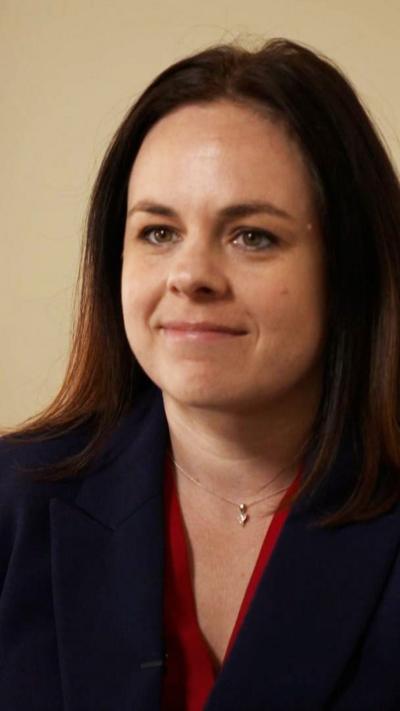 Kate Forbes - a young woman with long dark hair, seated and wearing a dark blazer and red blouse 