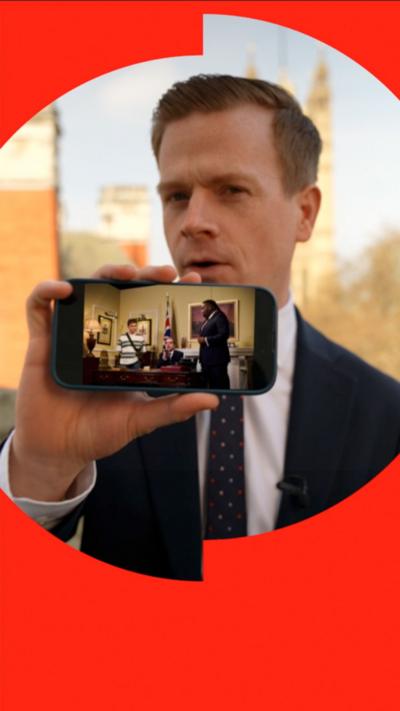 Nick Eardley holds a phone up with a picture of and SNL UK scene 