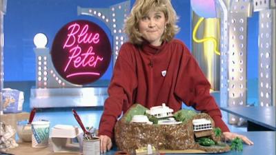 Anthea Turner in the Blue Peter studio.  Behind her are a few pieces of set including a 'Blue Peter' sign.  In front of her on the counter is a model of Tracy Island.  To the right are some of the materials she has used to make it, including yogurt posts, a bag of flour, brushes and mixing bowl.  She is wear a red jumper with a Blue Peter badge on.