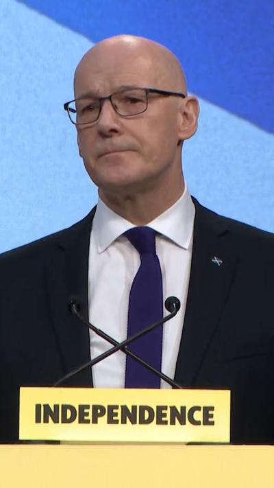 John Swinney stands at a conference podium, which has 'independence' in black letters on a yellow background