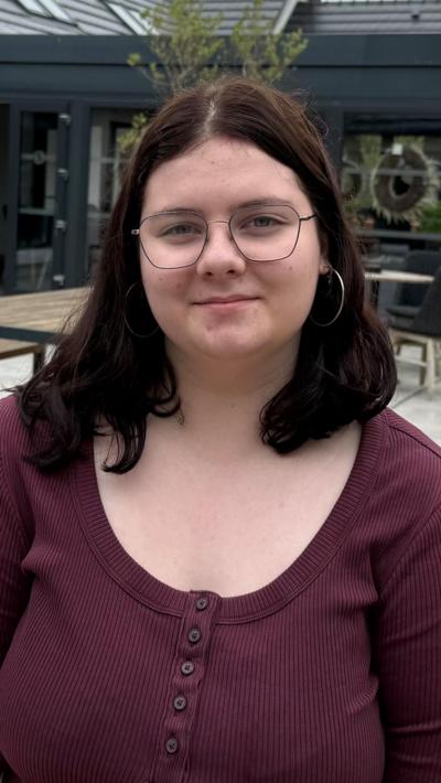 Aoísha is a young girl with shoulder-length dark hair. She is wearing a pair of wire glasses and large silver hoops. She is wearing a burgundy, ribbed shirt with buttons down the middle.