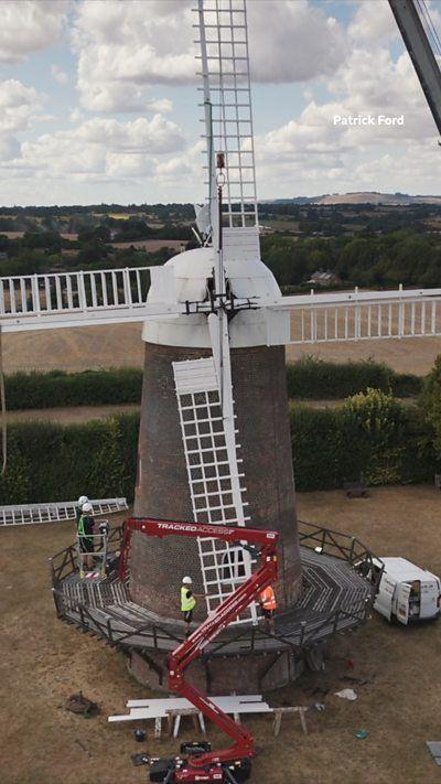 Wilton Windmill, a brick building with a white top and white sails. A cherry picker crane is in the foreground, with men in the basket moving towards the windmill.
