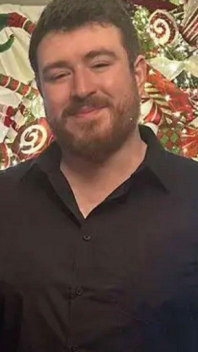 A picture of Zak Hughes. He is smiling at the camera. He is wearing a black, button up shirt and has dark hair and facial hair.