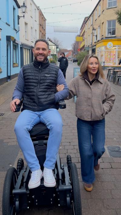 Phil Eaglesham in his wheelchair wearing blue jeans and travelling down the street with his wife. 