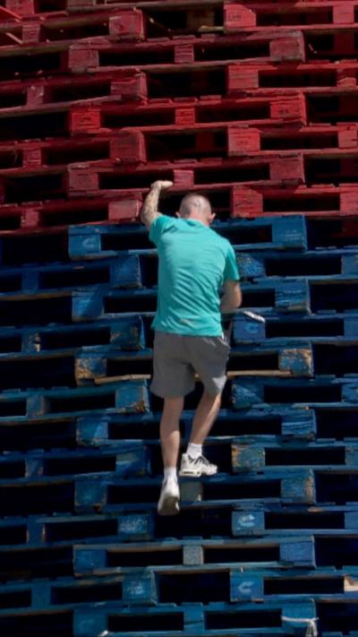 a man climbing a tack of red and blue pallets - he is wearing a T shirt, shorts and trainers