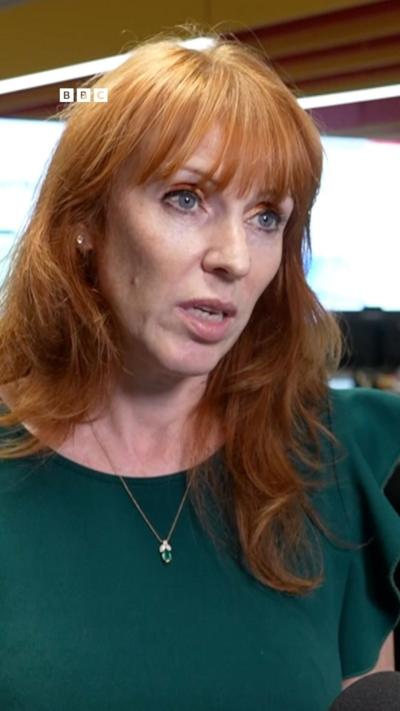 A woman with red hair looking to the right of camera wears a green blouse and a necklace.