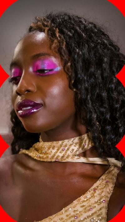 A woman with long, dark curly hair wears pink glittery eyeshadow and purple glittery lipgloss