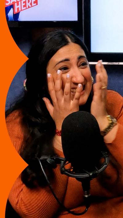 A woman wearing an orange jumper smiles and holds her hands up to her face.