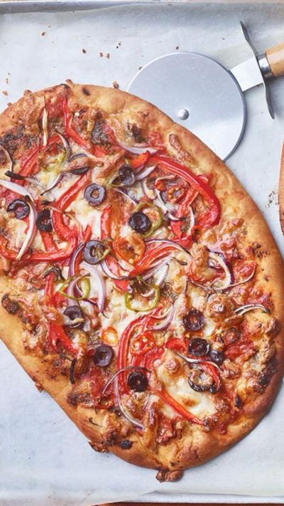 Oval shaped pizza with red peppers and olives, on a sheet of baking paper with a pizza cutter top right.