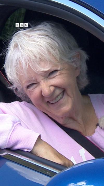 A woman wearing a pink top leans out of a car window.