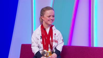 Paralympian Maisie Summers-Newton sits on the Newsround sofa with her two golds and one bronze medals, wearing her ParalympicsGB outfit