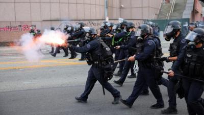 A line of riot police fire weapons as they advance down a street.