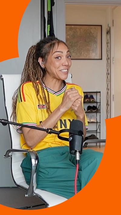 A woman is sat on a white chair with a mic on a stand in front of her. She's wearing a yellow T-shirt and green trousers.