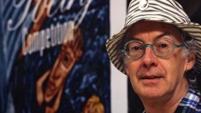 Roger McGough wears a striped hat. There is a competition poster in the background