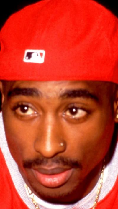 Tupac wears a red shirt and red baseball cap backwards. His left nostril has a piercing.