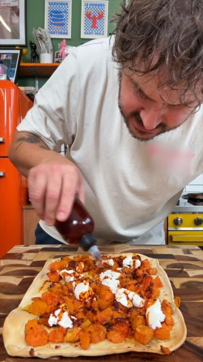 A man with short hair leans over a flatbread and pours oil over it