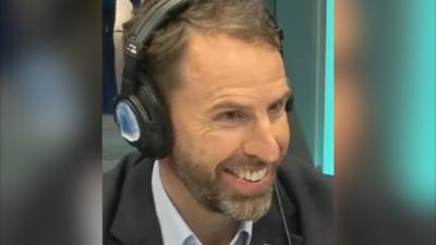 A man, Gareth Southgate, wears a shirt and suit jacket. He has headphones on and is smiling.