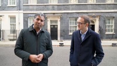 Two male journalists stand outside 10 Downing Street. One wears a dark green jacket and the other wears glasses and a navy jacket.