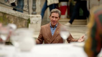 Rob Brydon opens his arms while singing to a table of people. He wears a brown suit