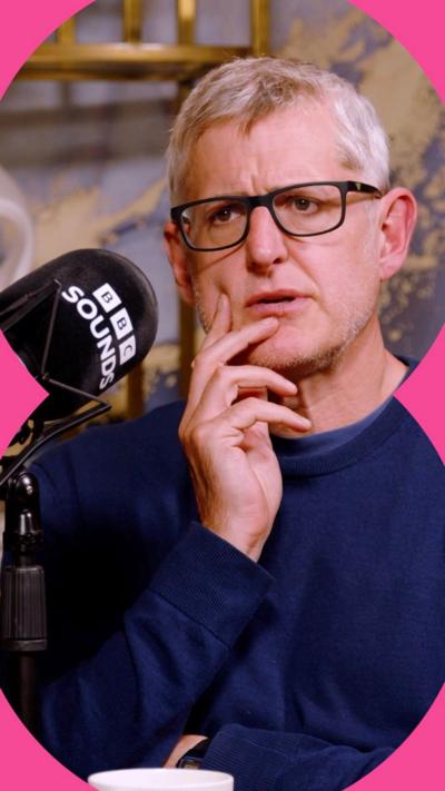 Louis Theroux sits with his hand on his pace looking confused. He wears a blue sweater.